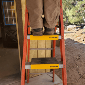 A person is standing the Rack-A-Tiers StepDek attached to an a-frame ladder.