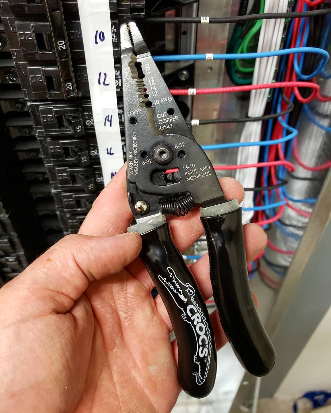 An electrician holding a pair of Croc’s Needle Nose Wire Strippers in front of a breaker panel.