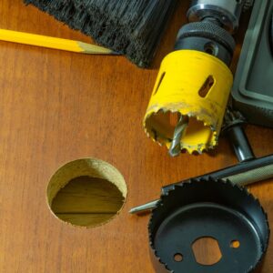 A drill with a hole saw bit has cut a circular hole in a piece of wood.