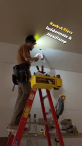 an electrician is standing on a ladder and wiring a light fixture. text reads: "rack-a-tiers ladder mate and headlamp"