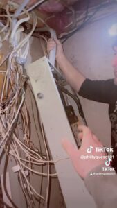 an electrician is removing an electrical panel from the wall in a residential basement. behind and around the panel are messy wires.