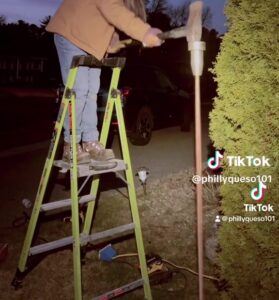 an electrician apprentice is striking a Rack-A-Tiers Sluggo-OX ground rod driver with the back of an axe. The electrician is standing on a yellow step ladder. they are outside and it is night.